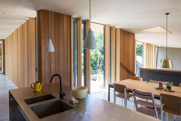 A bright, open-plan kitchen and dining area with extensive vertical timber wall panelling and full-height glazing. The kitchen island has a dark stone benchtop, integrated double sink, matte black gooseneck tap, and pendant lights. A timber dining table with upholstered chairs sits beyond the island, while a stair and dark balustrade are visible in the background. The space has a warm, contemporary timber-lined interior with strong natural light.