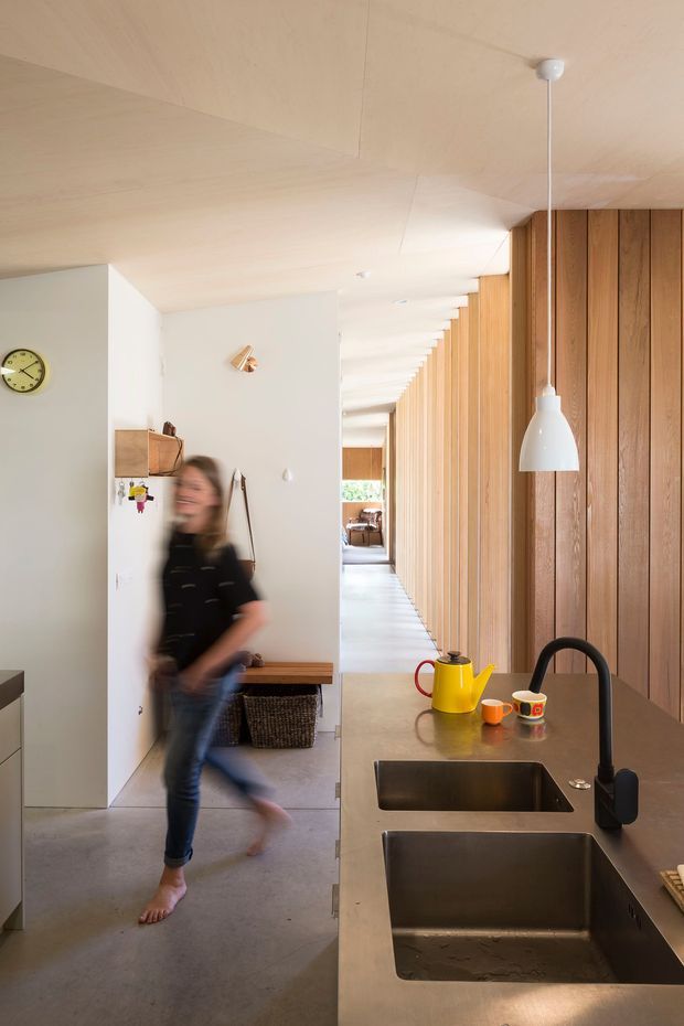 A bright, narrow open-plan kitchen and circulation space with a long double-bowl island in the foreground. The kitchen features warm vertical timber wall panelling, a neutral stone or concrete-look benchtop, dark undermount sinks, a matte black gooseneck tap, and a simple white pendant light. A white refrigerator, bench seat, woven basket, wall clock, and hallway are visible beyond, with polished concrete flooring and pale timber lining creating a warm, minimalist interior.