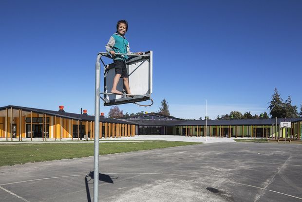 Exterior view of a low-rise school or community building beside an outdoor basketball court. The long building has dark horizontal roof forms, extensive glazing, and a warm timber-toned vertical facade screen, with a child standing on the basketball hoop structure in the foreground.