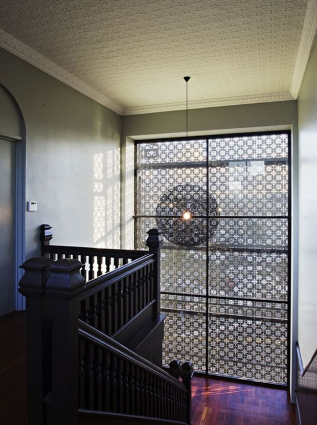 A grand interior stairway and landing with dark painted timber balustrades, polished dark timber flooring, pale walls, ornate ceiling cornices, and a textured patterned ceiling. A large floor-to-ceiling window with decorative geometric screening dominates the landing, casting patterned light into the space. The stair hall has a refined heritage character with strong contrast between the dark joinery and light finishes.