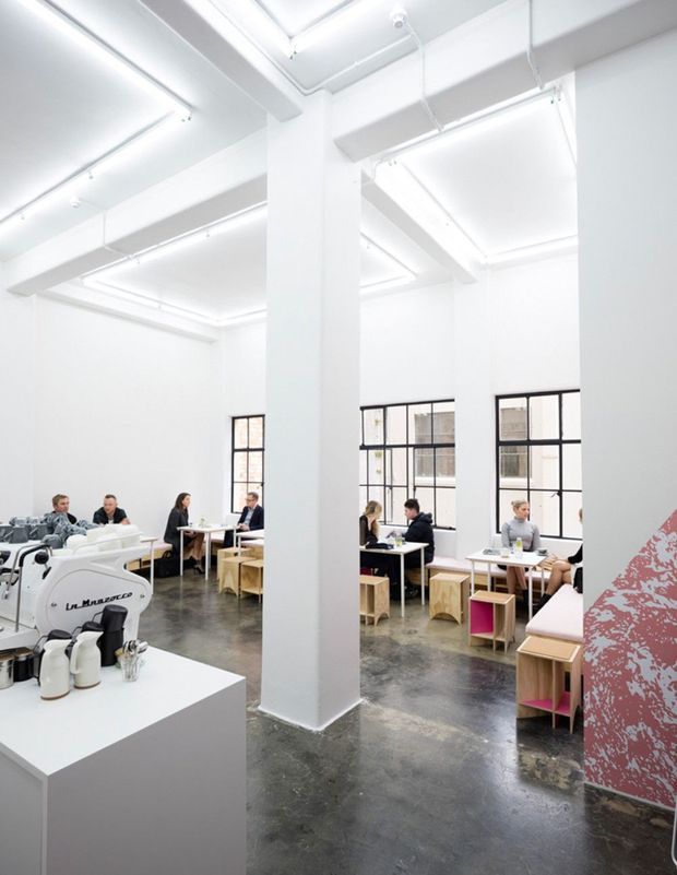 Bright, minimalist commercial café or hospitality interior with white walls, exposed structural columns, high ceilings, suspended linear lighting, large black-framed industrial-style windows, and a polished dark concrete floor. Several small tables and stools are arranged around a central coffee counter with a visible commercial espresso machine. The space has an open, gallery-like layout and a bold pink graphic feature panel at the right edge.
