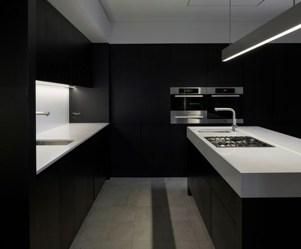 A sleek, contemporary galley kitchen in a predominantly black and white palette. Matte-black full-height cabinetry lines both sides, with integrated ovens, white benchtops, a gas cooktop, undermount sink, and a minimalist linear pendant light. Large grey floor tiles and concealed lighting create a refined, dramatic atmosphere.