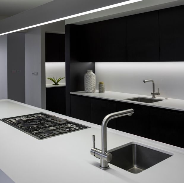 Contemporary minimalist kitchen with matte black full-height cabinetry, white solid-surface benchtops, integrated under-cabinet lighting, stainless-steel sinks and taps, and a large gas cooktop island. The space has clean linear detailing, concealed storage, and a high-contrast black-and-white palette.