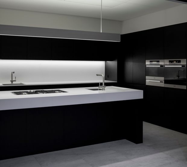 A sleek contemporary kitchen with extensive matte-black cabinetry, a large white island with an integrated gas cooktop and undermount sink, stainless steel mixer taps, built-in stainless appliances, a white illuminated splashback, and large grey floor tiles. The design has strong monochrome contrast and concealed storage.
