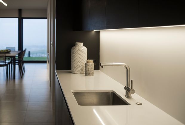 Contemporary kitchen interior focused on a long pale stone benchtop with an inset stainless-steel sink and curved chrome tap. Dark matte cabinetry with integrated under-cabinet lighting contrasts with the light work surface. A dining area and large glazed opening are visible beyond the kitchen.