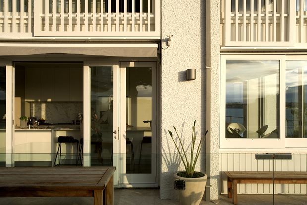 Exterior-facing view of a contemporary residential patio or terrace, with a textured light stucco facade, large glazed doors opening into a kitchen, an upper-level balcony screened by vertical slats, and a built-in bench beneath a window. A timber outdoor table and potted plant occupy the foreground. The warm low-angle sunlight creates a calm, inviting atmosphere.