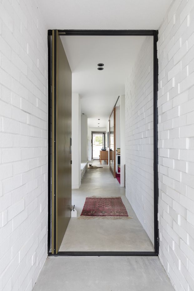 View through a dark-framed entry door into a long, bright residential hallway. White-painted brick walls and a pale concrete-like floor create a minimalist interior, with recessed ceiling lights, timber joinery and doors, and a patterned runner adding warmth. A glimpse of an open living area and rear glazing is visible at the far end.