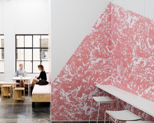 Contemporary commercial interior with a striking pink and white distressed graphic wall finish. A long white counter with stools runs along the feature wall, while a small café-style seating area with tables and benches sits beside large black-framed industrial-style windows. The polished dark floor and abundant daylight create a bold, bright workspace or hospitality setting.