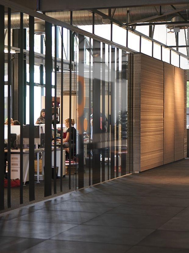 Interior view of a contemporary office or commercial workspace with a polished industrial character. The foreground is a broad dark tiled or concrete-finished circulation floor, while the workspace is enclosed by full-height black-framed glass partitions with vertical mullions. Exposed steel beams, services, and ductwork are visible overhead. A large fixed timber slatted wall or joinery volume forms a warm feature surface on the right, contrasting with the dark metal and glass. Desks and office workers are visible behind the glazing.