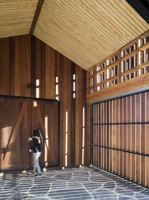 A tall, timber-lined interior space with a dramatic pitched ceiling, exposed dark timber framing, vertical timber wall boards, and a large slatted timber screen or garage-style door. Strong sunlight enters through narrow openings, creating striped shadows across the dark tiled or paved floor. A person stands beside a large timber door, emphasizing the generous vertical scale and architectural character.