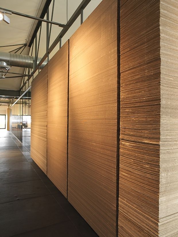 A long, narrow commercial or industrial interior corridor featuring a dramatic full-height partition wall clad in layered, textured warm-brown wood or wood-fibre panels. The space has dark polished flooring, exposed steel roof framing, visible ductwork, suspended track lighting, and glazed office or workspace areas beyond the partition. The composition emphasizes the repeating horizontal texture and warm illumination of the wall against the utilitarian exposed structure.