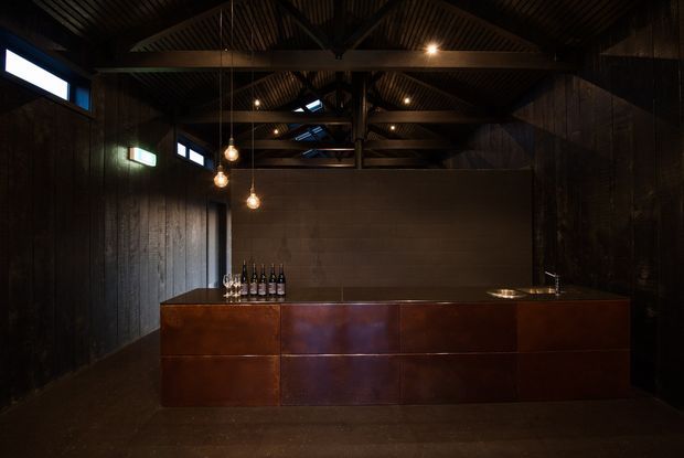 A dark, dramatic bar or entertaining space with a long copper-toned built-in bar counter and cabinetry, a small sink, and bottles displayed on the benchtop. The room has black-stained timber wall cladding, exposed dark timber roof trusses and corrugated ceiling panels, narrow clerestory windows, and multiple warm pendant lights. A concrete block partition wall forms the backdrop, creating an industrial and rustic atmosphere.