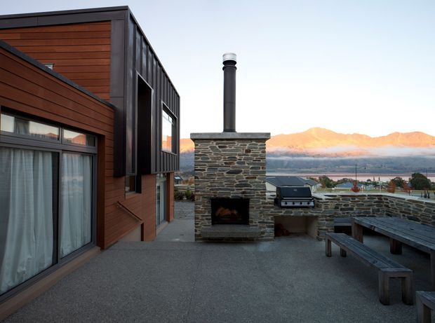 A contemporary outdoor terrace beside a dark metal and warm timber-clad dwelling. The space features a substantial rough-stone outdoor fireplace with chimney, integrated stone barbecue bench, and a dark timber dining table with benches. A lake and sunlit mountain range are visible beyond the terrace, creating a strong alpine lakeside setting.
