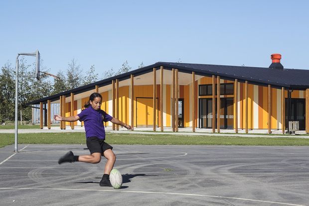 A low, elongated contemporary building borders an outdoor paved sports court. The building has a dark low-pitched roof, vivid yellow-orange walls, extensive glazing, and a regular rhythm of slender timber-coloured vertical posts and projecting structural elements. A child is playing soccer in the foreground, with open grass and trees beyond the building.