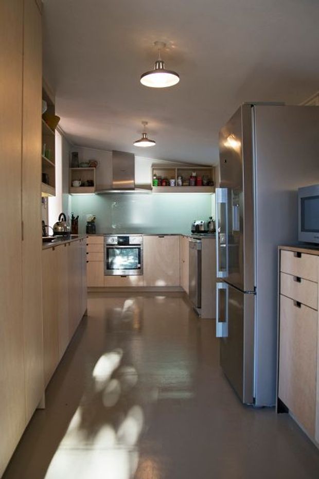 A compact galley-style kitchen with light timber cabinetry, stainless-steel appliances, a pale glass splashback, and integrated open shelving. The narrow space has a smooth reflective floor, simple pendant lighting, and a contemporary functional layout. A refrigerator and television cabinet are visible in the foreground.
