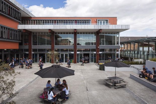 Wide exterior view of a contemporary multi-storey institutional or commercial building opening onto a large paved courtyard. The building features red-orange horizontal cladding, extensive floor-to-ceiling glazing, dark structural columns, projecting balconies with metal balustrades, and covered walkways. Students and outdoor seating with umbrellas occupy the courtyard, while additional covered structures are visible to the right.
