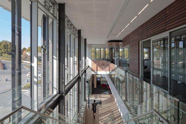 A bright multi-level interior circulation space in a contemporary commercial or institutional building. The view looks along an upper-level glazed walkway beside a stair descending to a lower level. Full-height windows line the left side, while the right wall combines dark horizontal timber-style cladding with extensive glazed doors and partitions. Glass balustrades, exposed structural columns, linear recessed lighting, and strong daylight create an airy atrium-like environment.