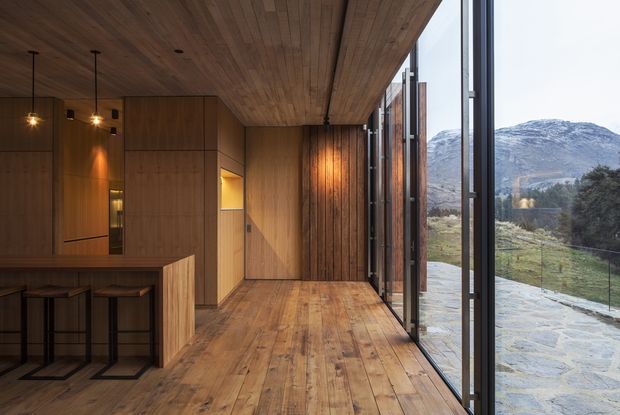 A warm, contemporary timber-lined interior with a compact kitchen and island seating in the foreground, opening through full-height glazed sliding doors to a dramatic mountainous landscape. Timber flooring, wall panelling, and ceiling create a cohesive cabin-like atmosphere, while slim dark window frames and minimal pendant lighting add contrast.