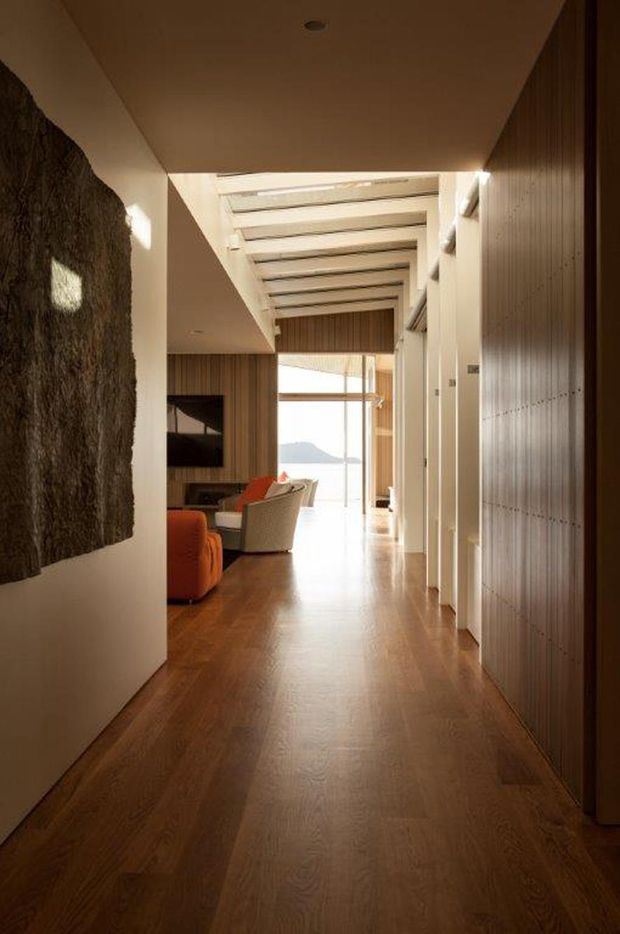 A warm, elongated interior hallway leads toward a bright living area and large glazed opening with distant landscape views. The corridor has rich timber flooring, extensive vertical timber wall panelling and joinery, a white ceiling with a linear skylight or clerestory structure, and recessed lighting. A dark textured feature artwork or wall surface occupies the left side, while a partial living room with orange and neutral seating is visible beyond.