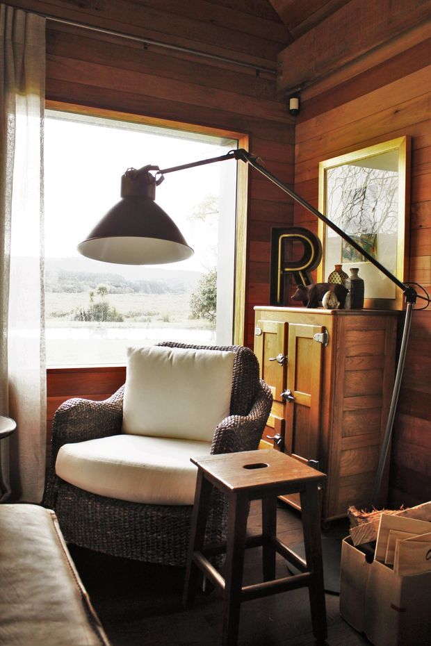 A cozy wood-panelled interior corner with a wicker armchair, small wooden side table, large black articulated floor lamp, and timber storage cabinet. A large window overlooks a rural landscape, while a framed mirror and decorative objects add visual depth. The space has a warm, rustic cabin-like atmosphere.
