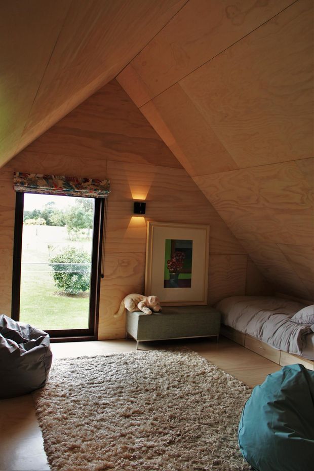A cozy attic-style bedroom with sloping timber-lined walls and ceiling, a large glazed door or window overlooking greenery, a low built-in bed, upholstered ottoman, framed artwork, beanbags, and a textured shag rug. The warm plywood interior and compact informal furnishings create a relaxed retreat-like atmosphere.
