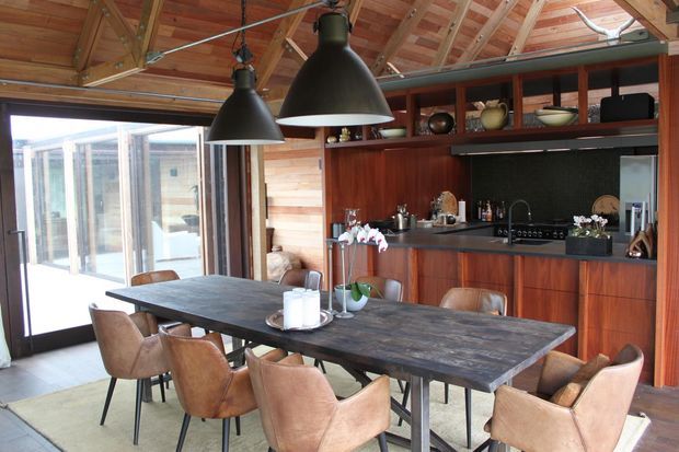 A warm, open-plan dining and kitchen interior with a large dark timber dining table surrounded by upholstered leather dining chairs. The kitchen features rich timber cabinetry, a dark benchtop, open display shelving, and black pendant lighting. Exposed timber roof framing and wall panelling create a rustic, lodge-like character, while extensive glazed doors connect the room to the outdoors.