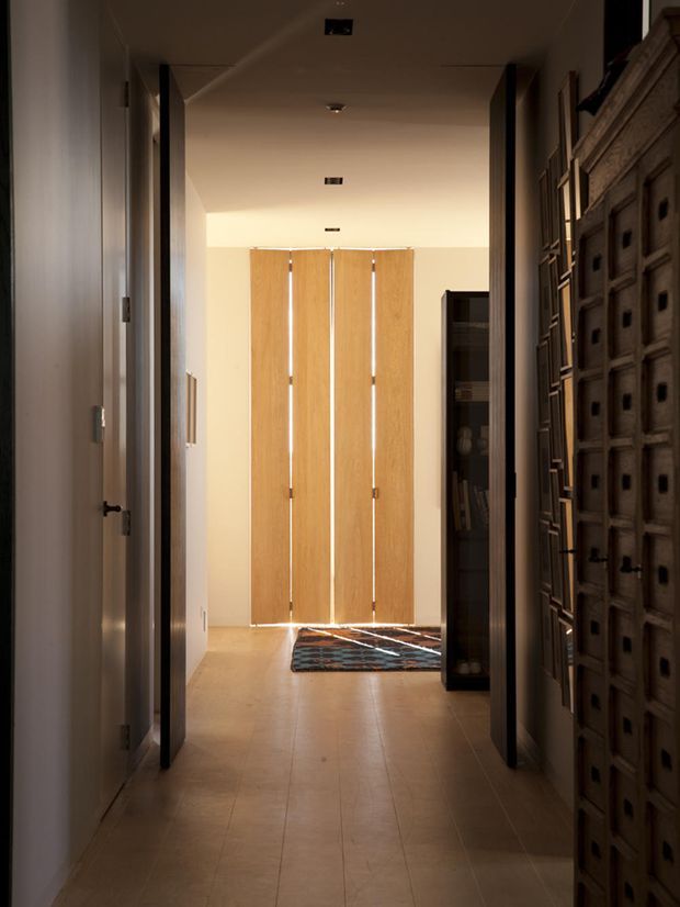 A warm, softly lit interior hallway looking toward tall timber-panelled doors or screens at the far end. The corridor has light tiled flooring, pale walls, dark door frames and recessed ceiling lights. A large distressed wooden drawer cabinet and open shelving line the right side, with a patterned rug near the far end.