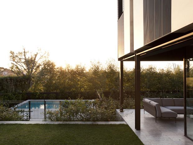 Contemporary residential exterior showing a covered outdoor living area with a large upholstered sofa, slim dark structural posts, extensive glazing, and pale rendered upper walls. The terrace overlooks a landscaped garden and swimming pool, with warm low sunlight filtering through trees.