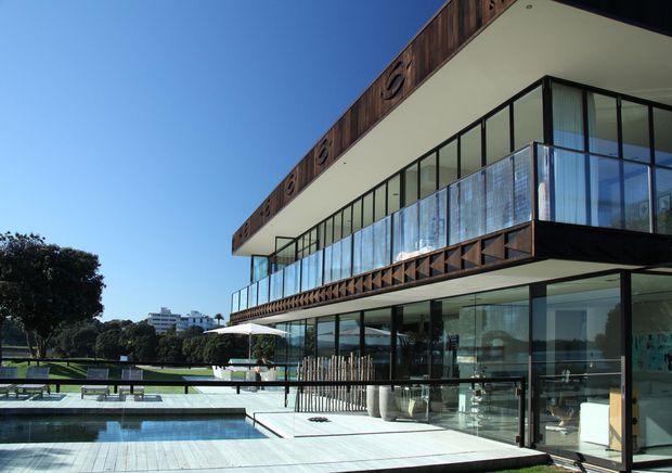 A modern multi-level residence with extensive floor-to-ceiling glazing, projecting balconies, dark timber fascia detailing, and a large pale deck surrounding a rectangular swimming pool. The house overlooks landscaped grounds and appears bright, open, and contemporary.