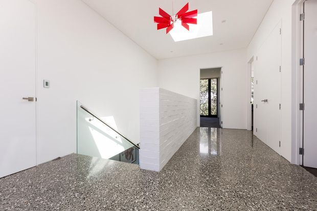 Bright modern hallway and stair landing with a polished exposed aggregate or terrazzo-like floor, white walls and flush white doors. A short white brick feature wall borders a glass stair balustrade, while a red sculptural pendant hangs below a rectangular skylight. Dark-framed glazing is visible at the far end.