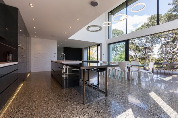 A spacious contemporary open-plan kitchen and dining area with a dark island, glossy black cabinetry, white textured masonry wall, polished terrazzo-style flooring, and extensive floor-to-ceiling glazing. A long dining table sits beside the windows, with sculptural circular pendant lights overhead and views of trees outside.