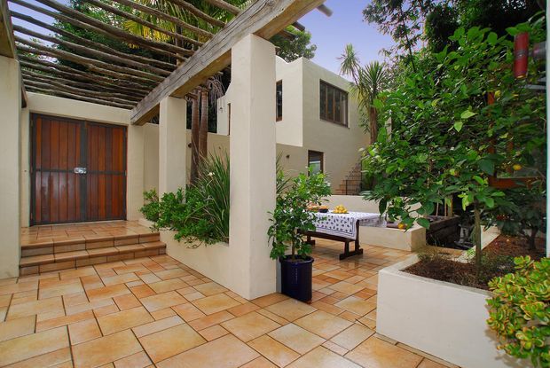 A warm Mediterranean-style courtyard entry with terracotta tiled paving, cream rendered walls, a timber garage or entry door, and a rustic timber pergola supported by square columns. Lush planting surrounds a small outdoor dining area with a covered table, creating a private garden courtyard.