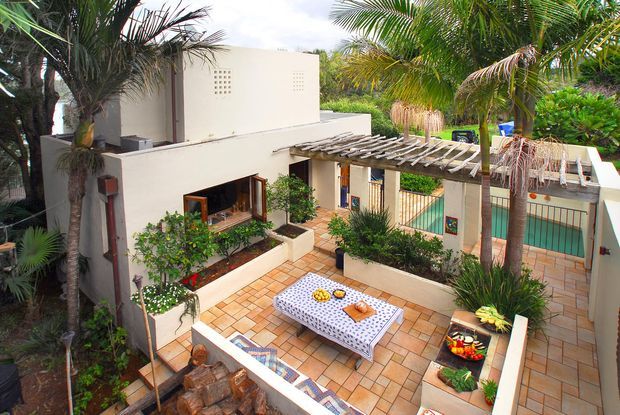 A sunlit tropical courtyard attached to a modern low-rise residence, featuring pale rendered walls, terracotta-toned tiled paving, raised planted garden beds, palm trees, and a timber pergola over a covered walkway. A central outdoor dining table and seating create an inviting entertaining area, with a turquoise pool or water feature visible beyond the courtyard.