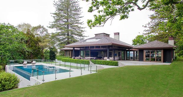 A single-storey pavilion-style residence set within a landscaped garden, featuring a broad hipped metal roof, dark timber-framed glazing, covered outdoor living areas, and a swimming pool with frameless glass balustrades in the foreground.