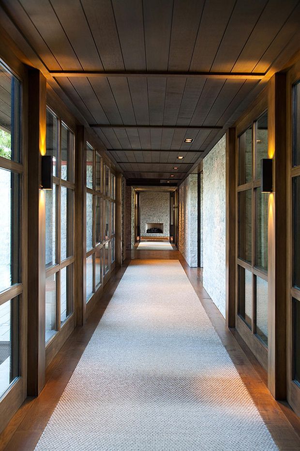 A long, refined interior hallway with extensive timber-framed glazing on both sides, dark timber wall and ceiling panelling, exposed textured stone walls, warm integrated wall lighting, and a pale carpet runner over timber flooring. The corridor leads toward a small fireplace or dark feature opening at the far end.