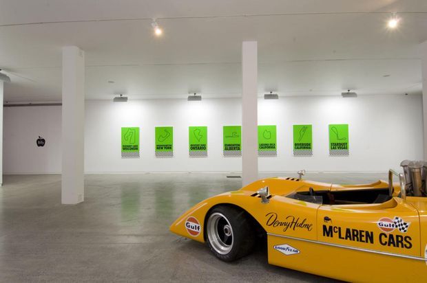 A spacious minimalist gallery or commercial exhibition interior with white walls, white structural columns, polished concrete flooring, track lighting, and a bright yellow McLaren race car displayed in the foreground. Green framed artworks or posters line the rear wall.