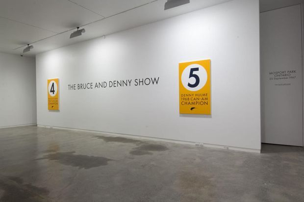 A minimalist contemporary gallery or museum exhibition space with white walls, polished concrete flooring, track lighting, and framed graphic posters reading “The Bruce and Denny Show” and “Denny Hulme 1968 Can-Am Champion.” A doorway at right is labelled “Mopar Park Sanitary,” reinforcing the exhibition or commercial gallery context.