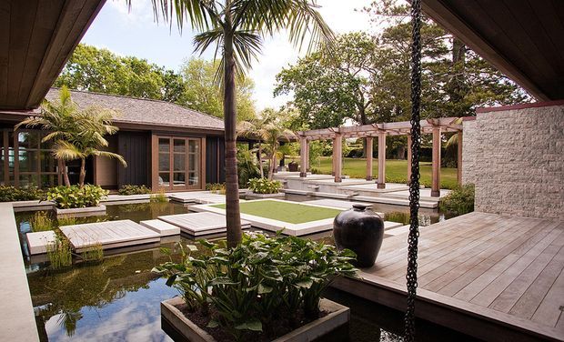 A landscaped courtyard-style outdoor living area with a shallow reflecting pond, timber decking, raised planters, tropical planting, and a timber-framed pavilion. The space includes a covered deck with a textured pale masonry feature wall, a large ceramic urn, and a pergola walkway surrounding a central lawn and water feature. The architecture has a resort-like, contemporary tropical character.
