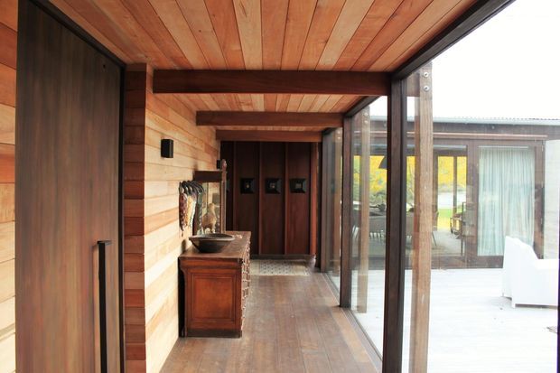 Warm timber-lined interior passage or entry hall with exposed ceiling beams, wide timber wall and floor boards, dark timber doors, and a traditional wooden console. Full-height black-framed glazing on the right opens visually toward a bright outdoor terrace and adjoining living area.