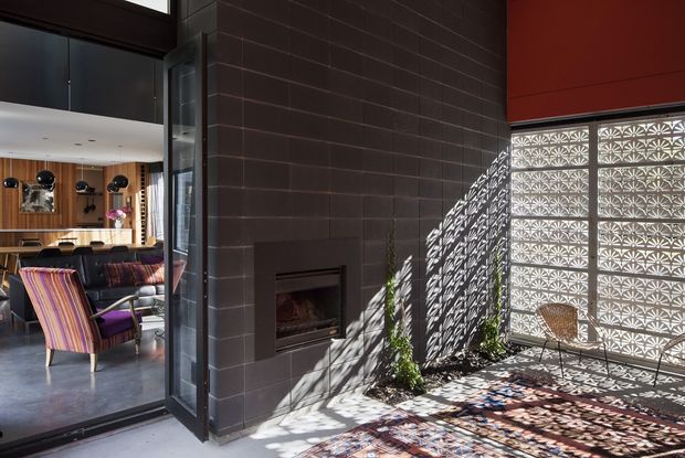 A contemporary indoor-outdoor living area centred on a dark tiled fireplace wall. The foreground features a partially enclosed patio or sunroom with patterned breeze-block screening, strong dappled sunlight, potted plants, and a woven chair. Through the open glazed doorway is a warm living room with timber wall panelling, lounge seating, and colourful upholstered armchairs.