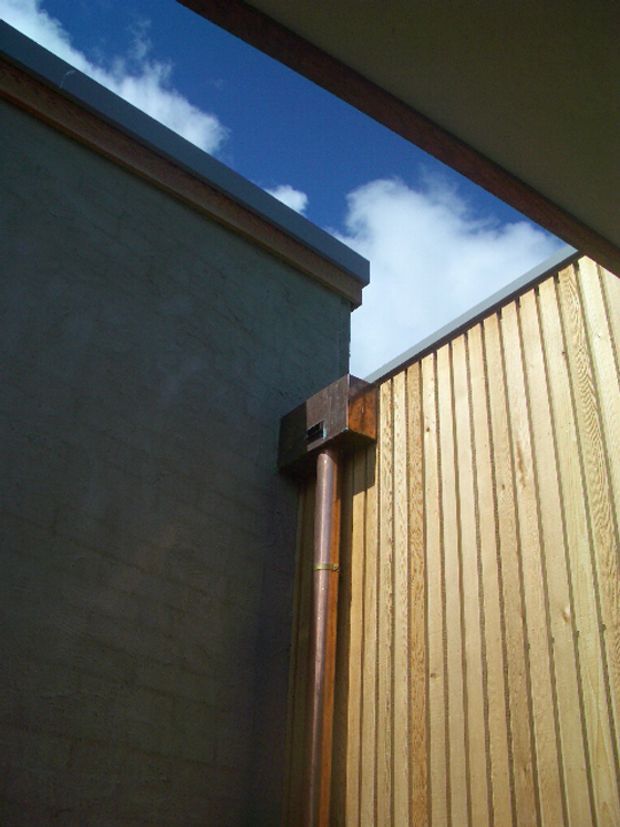 Close-up architectural detail of a narrow exterior courtyard or passage between a dark rendered masonry wall and a vertically ribbed timber-clad wall. A copper downpipe and metal roof-edge flashings are visible, with an open strip revealing blue sky and clouds above. The image emphasizes material junctions, drainage, and roofline detailing rather than a habitable room.