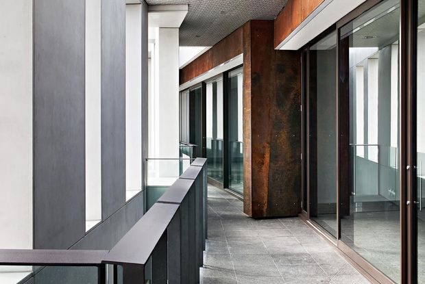 A modern covered balcony or external walkway with dark grey tiled flooring, full-height glazed doors and windows, glass balustrades, white structural columns, and a prominent rust-toned metal-clad wall or column. Reflections extend the corridor visually, while the linear composition and mixed materials create a refined contemporary architectural setting.