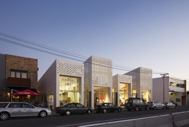 Street-level exterior view of a contemporary low-rise commercial retail building at dusk. The facade features several projecting white geometric volumes with perforated decorative screens, large illuminated shopfront glazing, and a contrasting exposed brown brick frontage. Multiple parked vehicles and overhead utility wires line the urban street.
