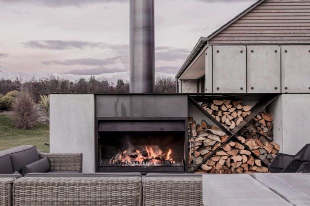 A contemporary outdoor entertaining area beside a timber-clad gabled building, featuring a built-in concrete or rendered outdoor fireplace with a tall metal chimney and neatly stacked firewood storage. Woven outdoor lounge furniture and a substantial pale stone or concrete table occupy the foreground, with a muted landscaped setting beyond.