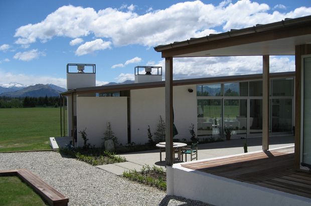 A low-slung modern house viewed from an outdoor terrace or courtyard. The building has crisp white rendered walls, extensive dark-framed glazing, a shallow roofline with rooftop vent structures, and a covered timber-decked outdoor seating area. Gravel landscaping, concrete paths, and sparse planting surround the house, with open fields and mountains visible beyond.