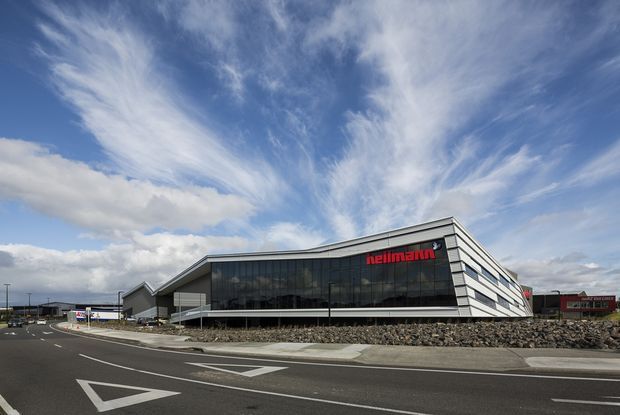 Wide exterior view of a large contemporary commercial building beside a paved roadway. The building has a dramatic angular, wedge-like form, extensive dark glass curtain walling, layered light-coloured horizontal facade bands, and a prominent red "hellmann" sign. A rock retaining wall and marked road occupy the foreground beneath a bright, cloud-filled sky.