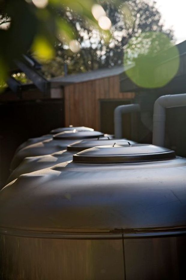 Close-up exterior view of several large cylindrical stainless-steel rainwater tanks with domed lids, connected by pale pipework. Timber fencing and leafy vegetation are visible in the background, with strong sun flare and shallow depth of field.