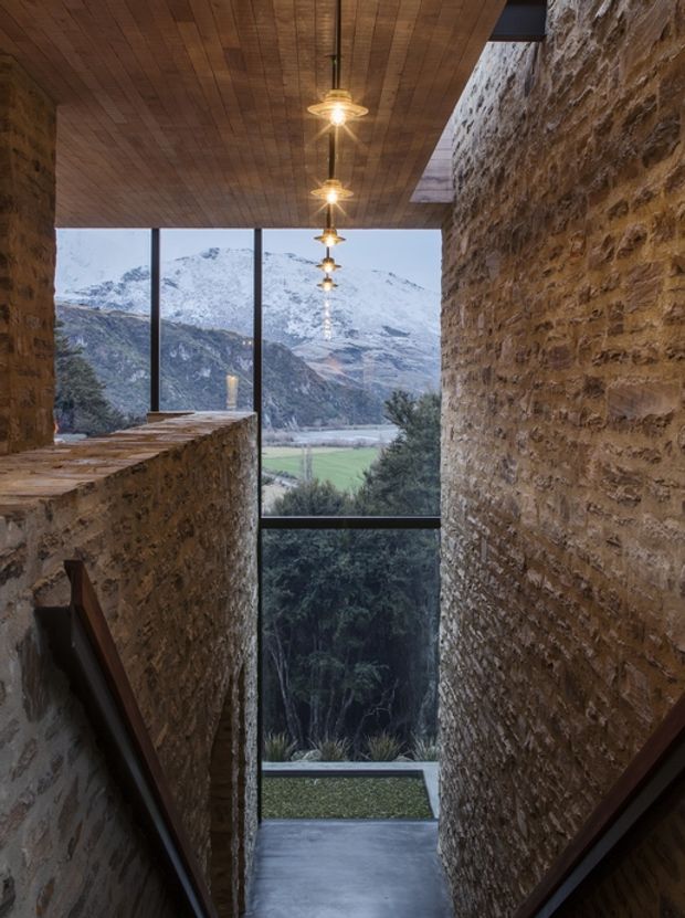 A narrow stairway or landing framed by rough exposed stone walls, with a warm timber-planked ceiling and a linear arrangement of pendant lights. Full-height glazing at the end opens to expansive views of a mountainous valley, fields, and trees. The space has a dramatic, rustic-modern character with dark stair rails, stone ledges, and a polished dark floor.