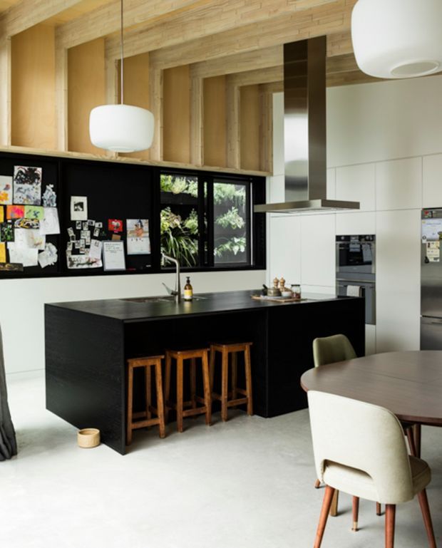 A contemporary open-plan kitchen and dining area with a large matte-black island, integrated sink, timber stools, white wall cabinetry, stainless-steel appliances, and a dark rangehood. Black-framed windows sit beneath a warm timber-lined ceiling structure, while a round dining table and upholstered chairs occupy the foreground. The room has a clean, modern look with strong black-and-white contrast softened by natural timber.