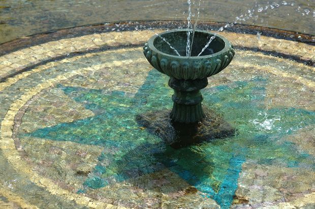 Close-up view of an ornate dark metal fountain bowl set at the centre of a shallow circular fountain basin. Water streams upward from the bowl and ripples across a multicoloured mosaic-tiled surface in muted beige, brown, turquoise, and blue tones.